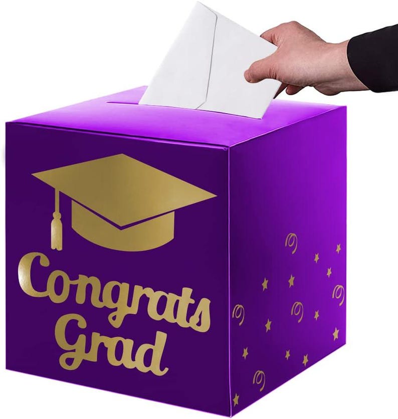 May include: A square purple card box featuring a gold graduation cap icon and the text "Congrats Grad" in gold lettering. The box has a slot on the top for inserting cards, and the sides are decorated with small gold stars and swirl patterns.