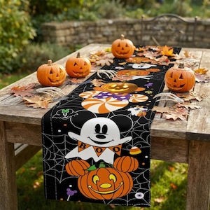 May include: A black table runner featuring a Halloween-themed pattern with white ghost characters, orange jack-o'-lanterns, spider webs, and candy corn. The runner displays the text BOO on a banner held by a ghost character with mouse ears.