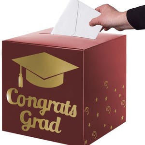 May include: A square, dark red cardboard box featuring a gold graduation cap icon and the text "Congrats Grad" in gold script. The side of the box is decorated with small gold stars and swirls. A white envelope is being inserted into the top slot.