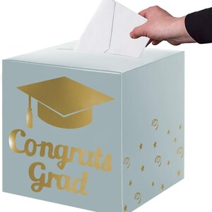 May include: A light blue square card box featuring a gold foil graduation cap icon and the text "Congrats Grad" on the front. The side panel is decorated with small gold foil stars and swirls, and there is a slot on the top for inserting cards.