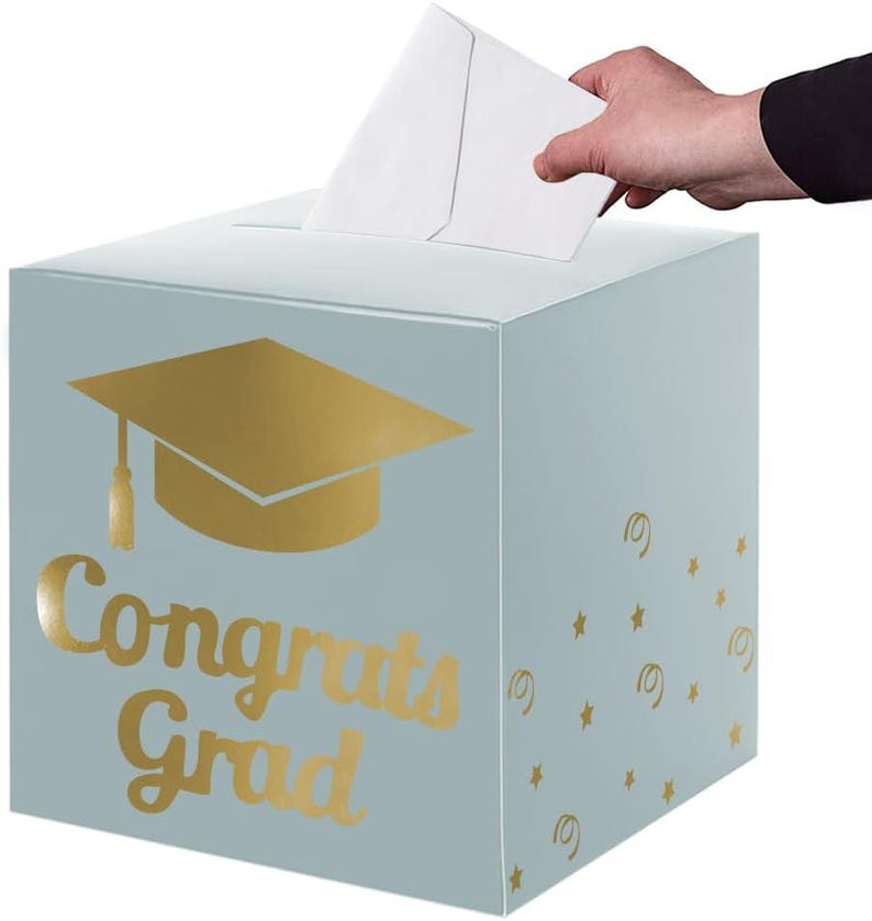 May include: A light blue square card box featuring a gold foil graduation cap icon and the text "Congrats Grad" on the front. The side panel is decorated with small gold foil stars and swirls, and there is a slot on the top for inserting cards.