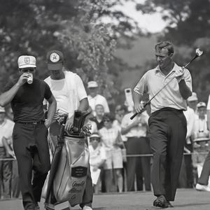 May include: Black and white photograph of a golf tournament. A golfer in a light-colored polo shirt and dark pants walks on the course, holding a golf club. Two men walk alongside him, one drinking from a cup, the other carrying a golf bag.