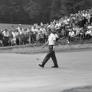 Puede incluir: Fotografía en blanco y negro de un golfista caminando por un campo de golf verde, sosteniendo un palo de golf. Una multitud de personas observa desde detrás de una colina cubierta de hierba. El golfista viste camisa blanca y pantalón oscuro.