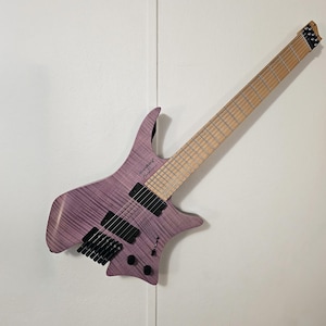 May include: A purple electric guitar with an unusual body shape and a light-coloured neck. The guitar has black pickups and tuning pegs. The brand name "strandberg" is visible on the body. The guitar is mounted on a white wall.