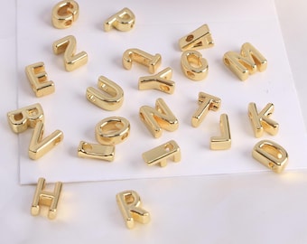Può includere: Una collezione di perline con lettere dell'alfabeto color oro. Le perline sono in vari caratteri e dimensioni, con fori per l'infilatura. Le lettere sono sparse su una superficie bianca, ideali per la creazione di gioielli o progetti di artigianato.