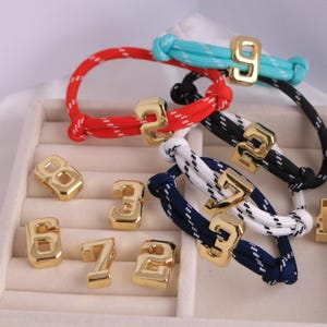 Pode incluir: Uma coleção de pulseiras de corda coloridas com pingentes de números dourados. As pulseiras são vermelhas, pretas, brancas, azuis e turquesas. Os números dourados 2, 3, 6, 7, 8 e 9 também estão presentes.