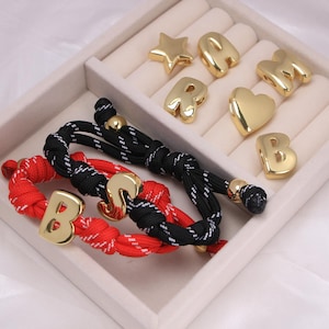 May include: A selection of gold-coloured charms, including letters, a star, and a heart, with two bracelets. One bracelet is red with a gold letter "B", and the other is black with a gold letter "S". The charms and bracelets are presented in a white jewellery box.