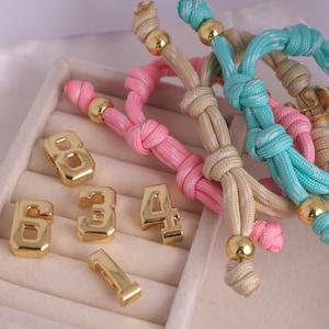 May include: Gold-colored metal number charms and colorful rope bracelets. The charms are numbers 1, 3, 4, 6, and 8. The bracelets are pink, beige, and blue with gold-colored beads. The bracelets are knotted.
