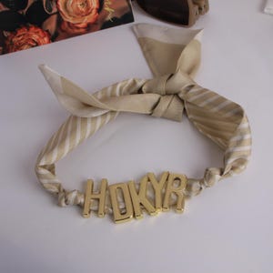 May include: A beige and white striped silk scarf tied in a knot, with gold-coloured letters spelling out "HDKYR" at the base. The scarf is a fashion accessory, likely for the hair or neck.