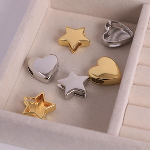 May include: A collection of heart and star-shaped charms in gold and silver tones. The charms are displayed in a light gray jewelry box. The charms have a hole for stringing onto a bracelet or necklace.