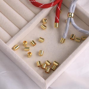 Könnte beinhalten: Nahaufnahme von goldfarbenen Metall-Schmuckzubehörteilen, darunter zylindrische Perlen und Kordelenden. Rote und graue geflochtene Kordeln mit goldenen Akzenten sind ebenfalls zu sehen. Die Teile sind in einer weißen, unterteilten Schale angeordnet.
