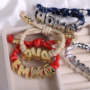 Puede incluir: Una colección de pulseras personalizadas con nombres y la palabra "HOME". Las pulseras presentan letras de metal dorado y plateado, y bandas de estilo cuerda en rojo, blanco, azul marino y beige. Las pulseras se exhiben sobre una superficie clara.