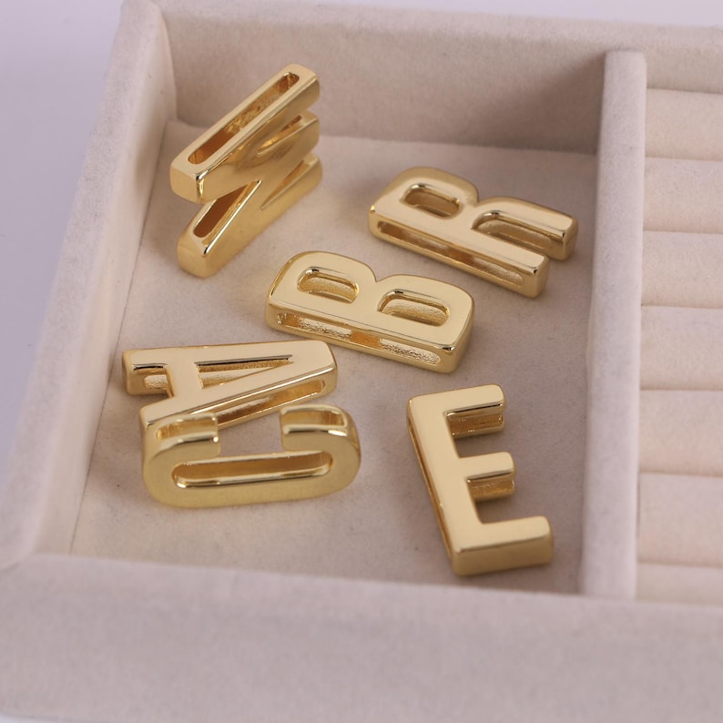 May include: A selection of gold-coloured metal letter charms, including A, B, E, and W, are presented in a light-coloured jewellery tray. The block-style letters have a shiny finish, suitable for jewellery making or crafting.