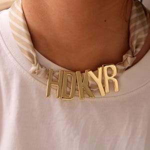 May include: A gold-coloured necklace spelling "HDKYR" worn with a striped scarf and a white t-shirt. The necklace is the main feature, with the letters clearly visible. The scarf adds a pattern and texture.