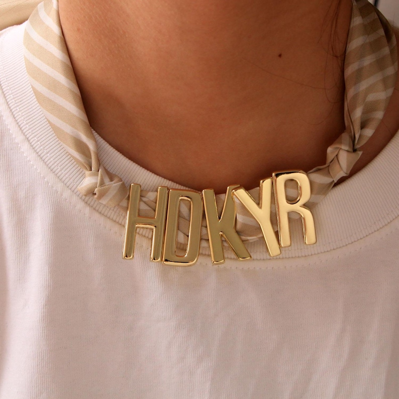 May include: A gold-coloured necklace spelling "HDKYR" worn with a striped scarf and a white t-shirt. The necklace is the main feature, with the letters clearly visible. The scarf adds a pattern and texture.