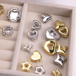 Puede incluir: Un surtido de cuentas met&aacute;licas plateadas y doradas en varias formas, incluyendo corazones, estrellas y tr&eacute;boles de cuatro hojas. Las cuentas est&aacute;n dispuestas en una bandeja dividida, mostrando sus diferentes dise&ntilde;os y acabados.