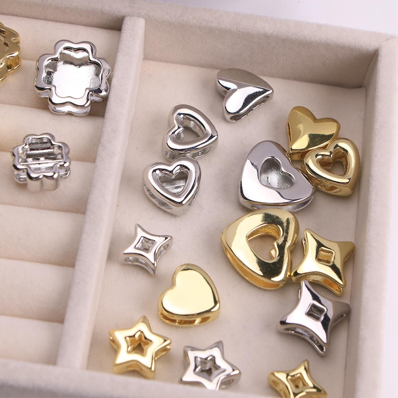 Puede incluir: Un surtido de cuentas met&aacute;licas plateadas y doradas en varias formas, incluyendo corazones, estrellas y tr&eacute;boles de cuatro hojas. Las cuentas est&aacute;n dispuestas en una bandeja dividida, mostrando sus diferentes dise&ntilde;os y acabados.