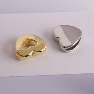 Puede incluir: Dos colgantes en forma de coraz&oacute;n. Uno es de color dorado, con un acabado pulido. El otro es de color plateado, tambi&eacute;n con un acabado pulido. Ambos colgantes tienen un dise&ntilde;o suave y redondeado y una peque&ntilde;a abertura lateral.