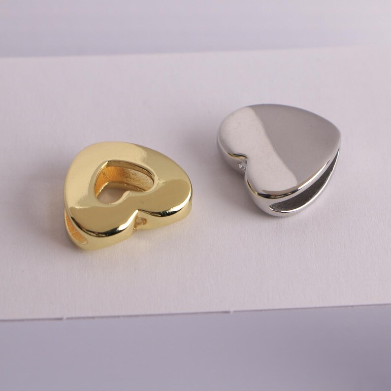 Puede incluir: Dos colgantes en forma de coraz&oacute;n. Uno es de color dorado, con un acabado pulido. El otro es de color plateado, tambi&eacute;n con un acabado pulido. Ambos colgantes tienen un dise&ntilde;o suave y redondeado y una peque&ntilde;a abertura lateral.