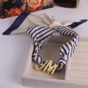 May include: A cream silk scarf with navy blue stripes and trim. The scarf is knotted and features a gold metal pendant with the letters "JM". The scarf is displayed in a white jewellery box.