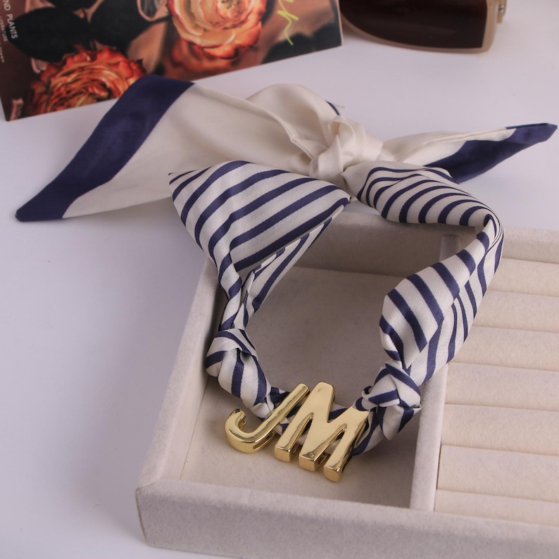 May include: A cream silk scarf with navy blue stripes and trim. The scarf is knotted and features a gold metal pendant with the letters "JM". The scarf is displayed in a white jewellery box.