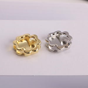 Puede incluir: Dos componentes met&aacute;licos en forma de flor, uno dorado y otro plateado, se muestran sobre una superficie blanca. Los componentes tienen un dise&ntilde;o en capas con una depresi&oacute;n central, adecuados para la creaci&oacute;n de joyas o proyectos de artesan&iacute;a.