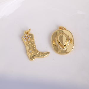 May include: Two gold-coloured charms: a cowboy boot and a cowboy hat. The boot charm features floral and crystal accents. The hat charm has a crystal-lined brim and a detailed centre. Both charms have a loop for attaching to a necklace or bracelet.