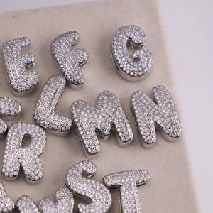 May include: Close-up of several silver-colored, gem-studded alphabet letters. The letters are in a bold, rounded font and are encrusted with small, sparkling gems. The letters are arranged on a light-colored surface.