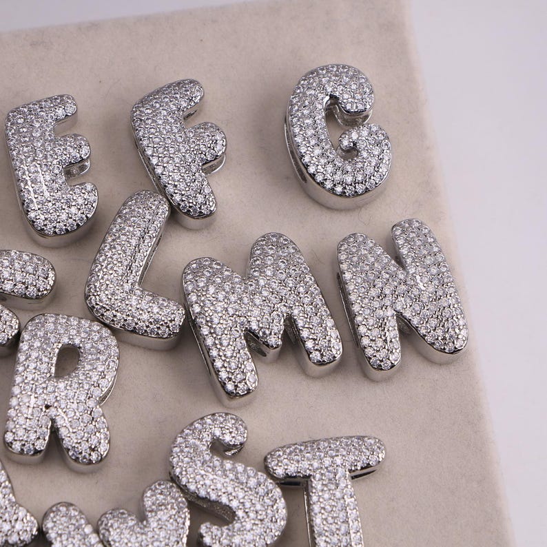 May include: Close-up of several silver-colored, gem-studded alphabet letters. The letters are in a bold, rounded font and are encrusted with small, sparkling gems. The letters are arranged on a light-colored surface.