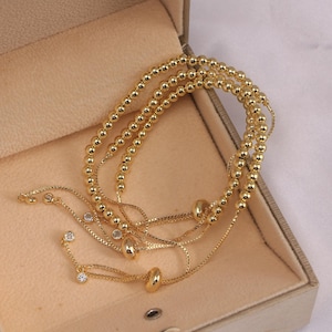 May include: Gold-coloured beaded bracelets and delicate chain bracelets with small, clear gemstones. The jewellery is displayed inside a beige jewellery box. The bracelets have a shiny, polished finish.