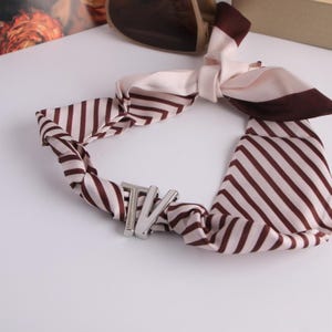 May include: A silk scarf with brown and cream stripes. It has a silver-coloured metal clasp with the letters "TV". The scarf is knotted, with the ends hanging down. The scarf is a stylish accessory.
