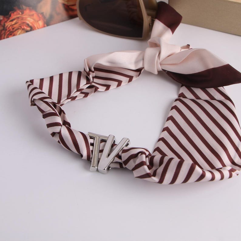 May include: A silk scarf with brown and cream stripes. It has a silver-coloured metal clasp with the letters "TV". The scarf is knotted, with the ends hanging down. The scarf is a stylish accessory.