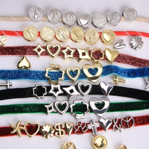 Puede incluir: Un surtido de abalorios y cuentas met&aacute;licas en diversas formas y dise&ntilde;os. La colecci&oacute;n incluye formas de coraz&oacute;n, estrella, cruz y flor, junto con medallones religiosos redondos. Los abalorios son en tonos dorados y plateados, dispuestos sobre cintas de colores.