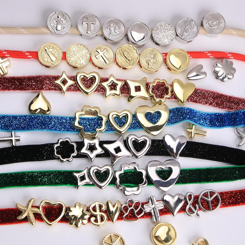 Puede incluir: Un surtido de abalorios y cuentas met&aacute;licas en diversas formas y dise&ntilde;os. La colecci&oacute;n incluye formas de coraz&oacute;n, estrella, cruz y flor, junto con medallones religiosos redondos. Los abalorios son en tonos dorados y plateados, dispuestos sobre cintas de colores.