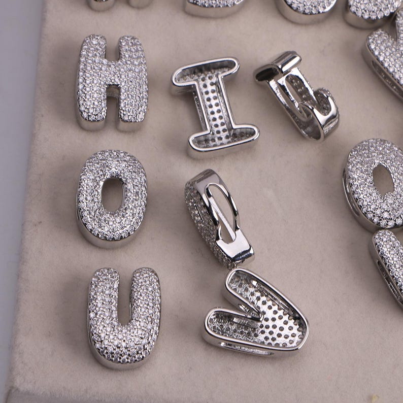 May include: Close-up of silver-toned letter pendants encrusted with small, clear gemstones. The letters include H, I, O, and U. The pendants are likely for jewelry making or personalization.