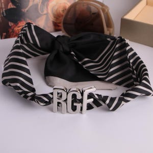 May include: A black and white striped scarf with a silver metal buckle spelling "RGF". The scarf is knotted in the middle and has a satin finish. Sunglasses and a box are in the background.