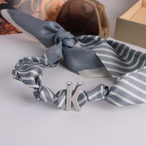 May include: A grey and white striped silk scarf with a silver letter "K" detail. The scarf is knotted and has a soft, flowing appearance. Sunglasses and a small box are in the background.