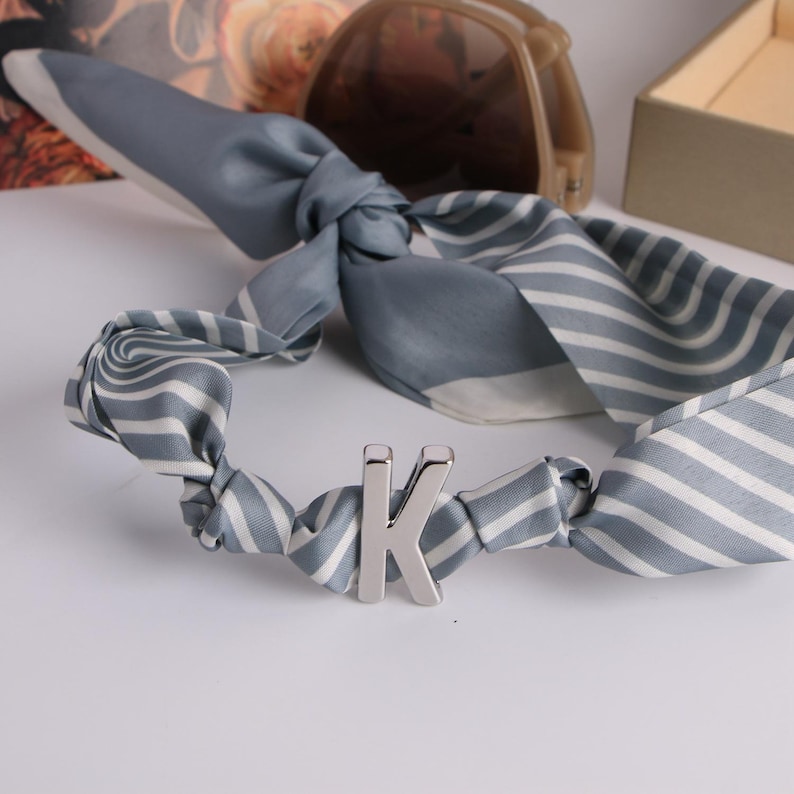 May include: A grey and white striped silk scarf with a silver letter "K" detail. The scarf is knotted and has a soft, flowing appearance. Sunglasses and a small box are in the background.