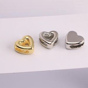 Puede incluir: Tres cuentas en forma de coraz&oacute;n en oro, plata y plata pulida. Cada cuenta tiene un centro hueco y un agujero para ensartar. Las cuentas se muestran sobre una superficie blanca.