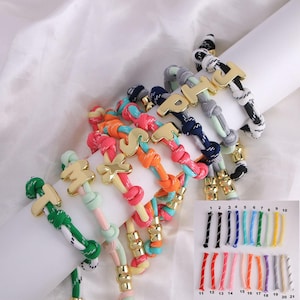 May include: A selection of rope bracelets in vibrant colours, each adorned with gold-tone letter charms. The bracelets display a variety of colour schemes, such as pink, orange, green, and blue. A colour chart with numbered rope samples is also present.