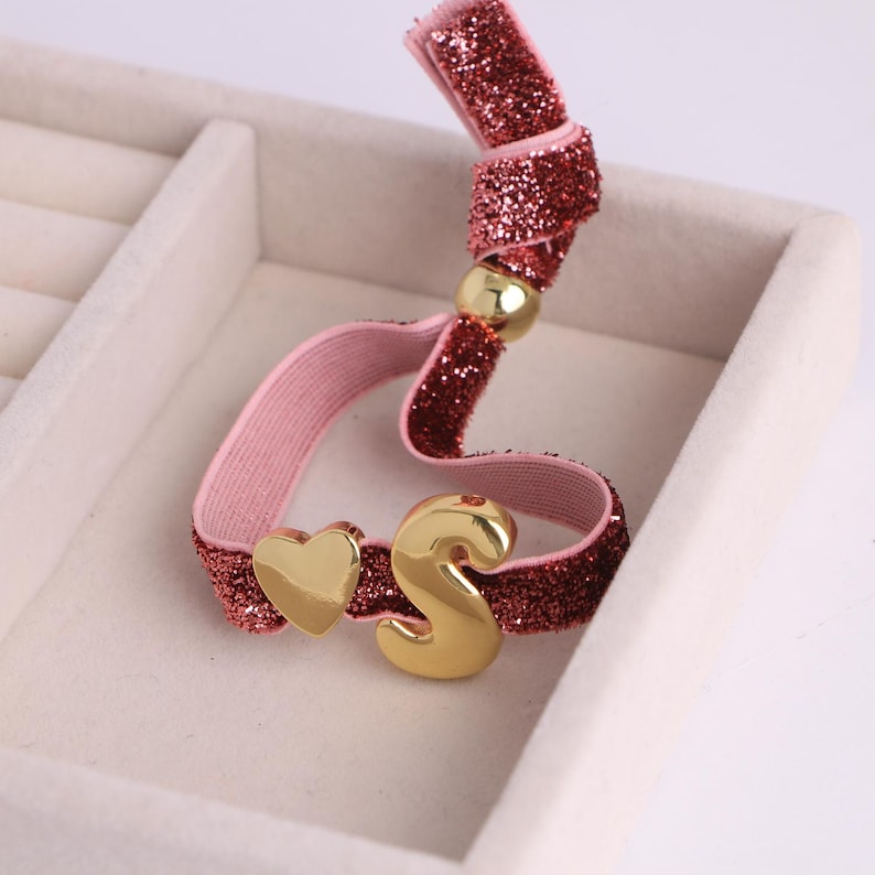 Puede incluir: Una goma para el pelo rosa y roja con purpurina, con un coraz&oacute;n dorado y la letra "S". La goma para el pelo tiene una cuenta dorada. El fondo es blanco.