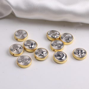 May include: A collection of round, gold-rimmed beads with silver-colored embossed designs. The designs include a lightning bolt, a heart, a star, a dollar sign, a crescent moon, and a four-leaf clover. The beads are scattered on a white surface.