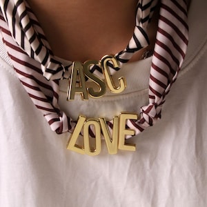 Może przedstawiać: Złoty metalowy naszyjnik z napisami "ASC" i "LOVE" noszony z paskowanym szalikiem. Szalik ma wzór czarnych, białych i bordowych pasków. Naszyjnik jest prezentowany na białym tle.