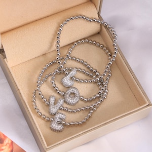 May include: Silver beaded bracelets spelling out the word "LOVE" with sparkling, clear stones. The bracelets are displayed inside a beige jewelry box, creating a romantic and elegant presentation.