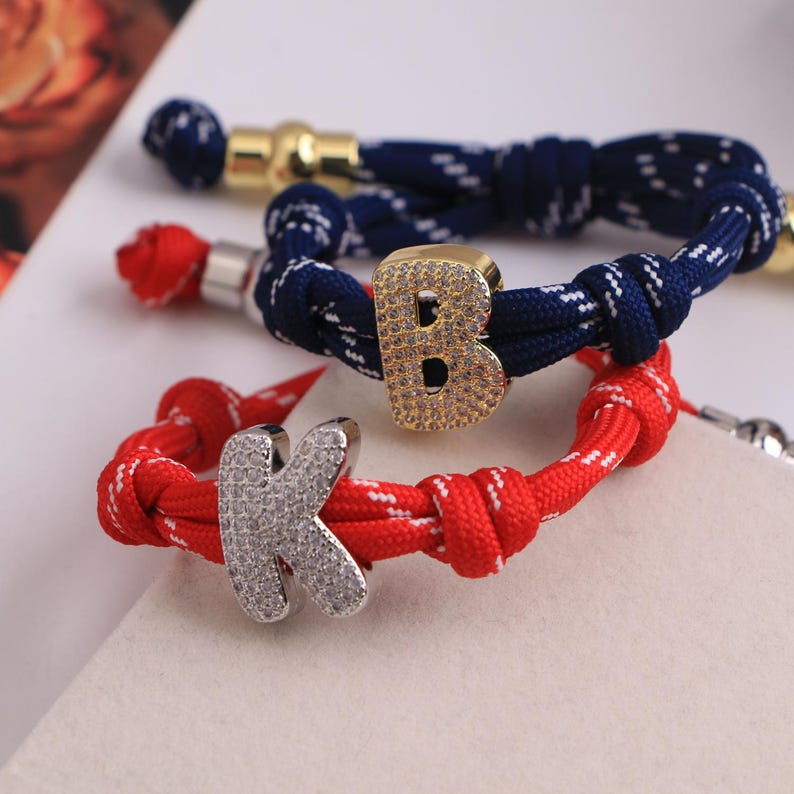 May include: Three colorful rope bracelets with letter charms. One bracelet is red with a silver 'K' charm, another is navy blue with a gold 'B' charm, and the third is navy blue with gold accents.