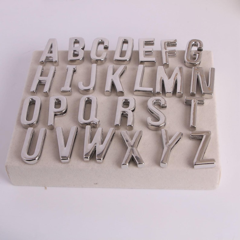 May include: A full set of silver metal alphabet letters, from A to Z, displayed on a light beige rectangular surface. The letters are in a bold, sans-serif font and are evenly spaced.