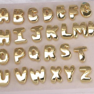 Op de afbeelding: Een verzameling goudkleurige alfabetbedels, elke letter in een afgerond, bubbelachtig lettertype. De letters zijn in rijen gerangschikt en tonen het hele Engelse alfabet van A tot Z. De bedels hebben een glanzend, reflecterend oppervlak.