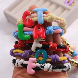 Op de afbeelding: Een collectie armbanden gemaakt van kleurrijke gevlochten nylon koorden, elk voorzien van een glanzende, met emaille gecoate letterbedel. De letters zijn uitgevoerd in diverse felle kleuren zoals rood, blauw, groen, wit, paars en roze, met goudkleurige randen.