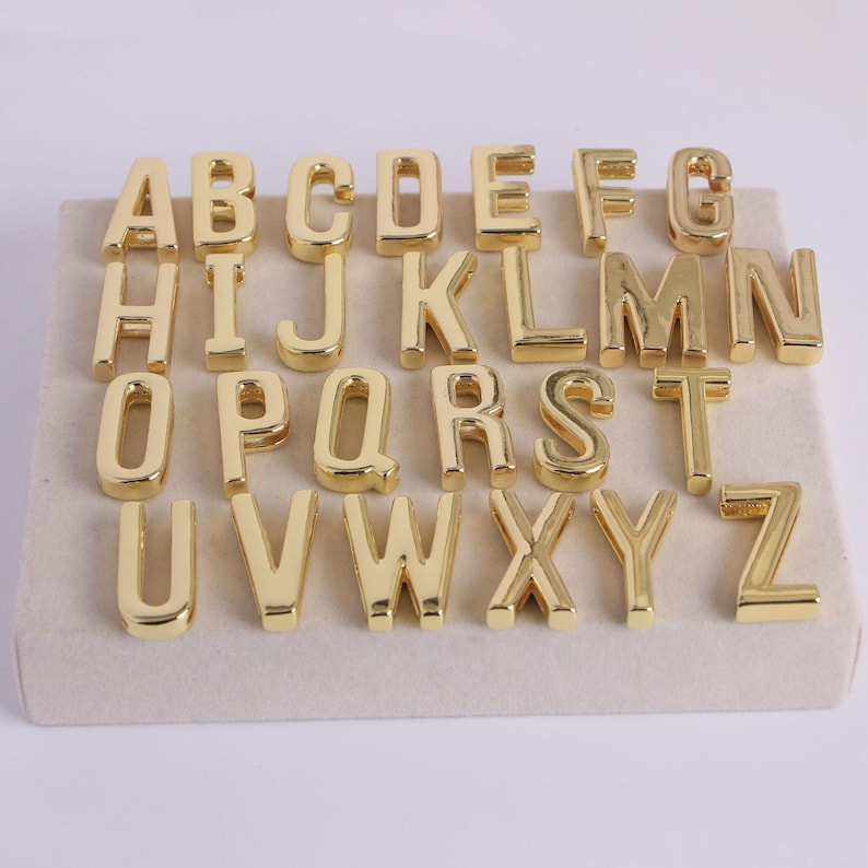 May include: Gold-coloured metal alphabet letters A to Z arranged on a beige surface. The letters are in a serif font and are evenly spaced. The letters are shiny and reflect light.