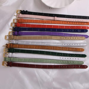Op de afbeelding: Een verzameling kleurrijke horlogebandjes in verschillende tinten, waaronder zwart, roze, oranje, rood, blauw, paars, goud, zilver, bruin, groen, wit en bordeaux. Elke band heeft een goudkleurige gesp en een textuur met krokodillenpatroon.